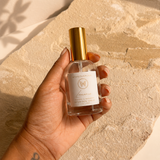 A hand holding a clear glass perfume bottle with a gold cap and a white label, set against a textured beige stone background with warm sunlight and leaf shadows.