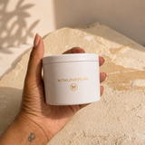 A hand holding a white cosmetic jar with gold lettering that reads 'WITH LOVE PEARL' and a gold monogram logo, set against a textured beige stone background with warm natural shadows.