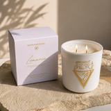 A white candle in a white glass jar with gold geometric branding sits beside its matching white box on a textured stone slab. The candle is lit with two visible flames, and the scene is styled in warm natural light with soft shadows.