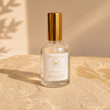 A clear glass spray bottle with a shiny gold cap and a white label reading 'WITH LOVE PEARLS' and 'Lumina', placed on a textured beige stone slab with warm sunlight and leaf shadows in the background.