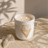 A white double-wick candle with a gold geometric logo sits on a textured stone slab with natural shadows cast across it. Two clear crystals rest beside the candle, and the background features a warm beige wall with leaf shadows.