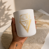A hand holds a white ceramic candle jar decorated with a gold geometric triangle design. The background features a textured beige stone slab and soft shadows cast by leafy branches.